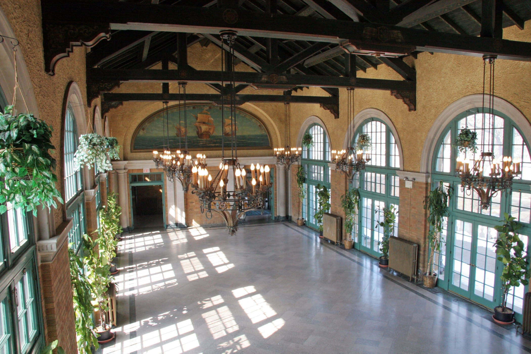 Grand hall with high ceilings, arched windows, and hanging chandeliers. A mural of ships adorns the back wall. Potted plants line the sides. Sunlight streams through the windows.